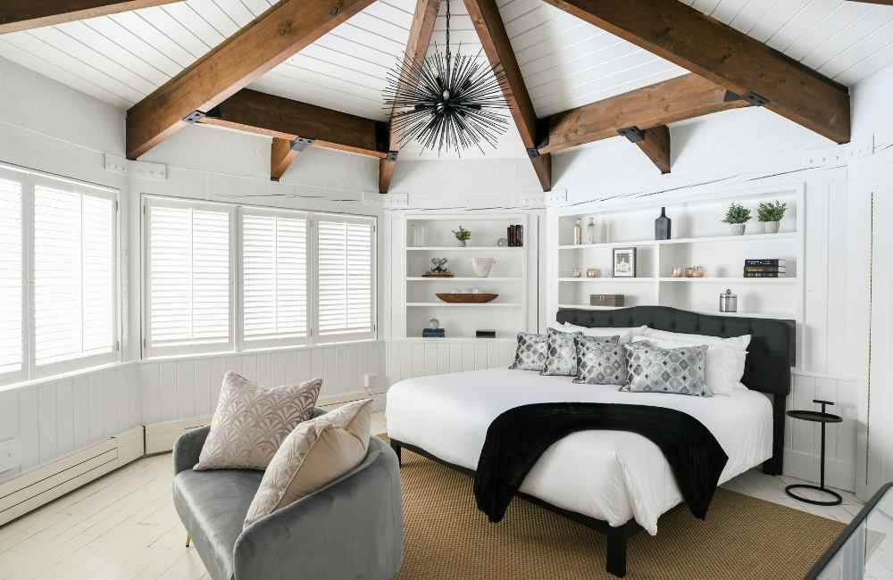 White bedroom with wooden beams and windows, white bedding on bed with decorative blanket.